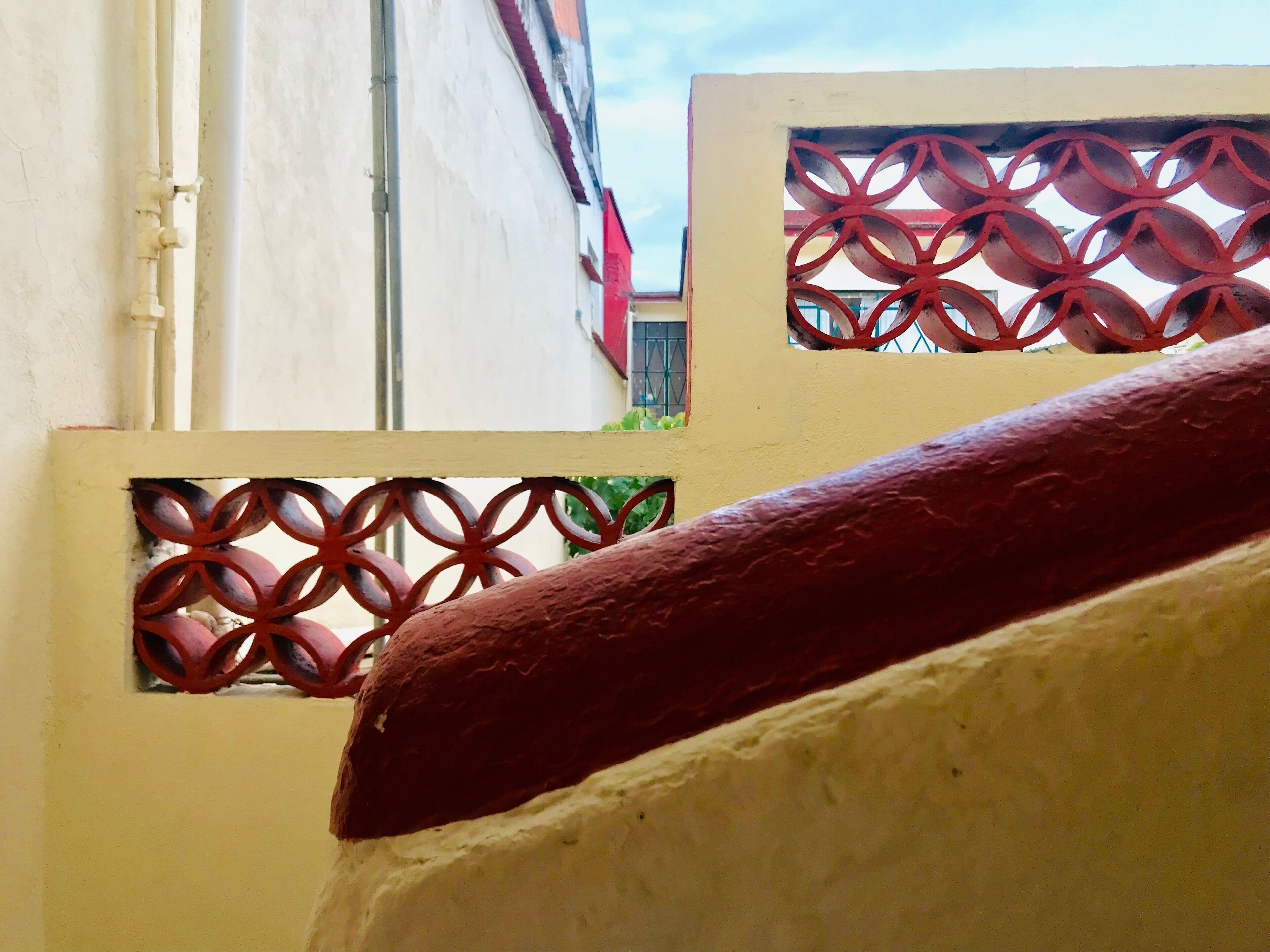 Une vue à travers un portail en métal décoratif orné de motifs géométriques, qui mène à un bâtiment doté d’un toit rouge et d’une fenêtre avec des barreaux en métal.