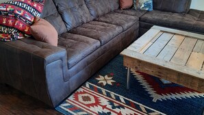 Een woonkamer met een bruine leren hoekbank, een houten salontafel, een kleurrijk vloerkleed en een witte bijzettafel met een lamp.