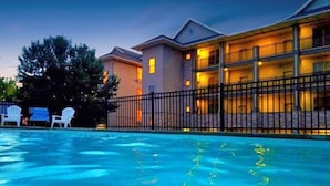 A swimming pool with clear water, surrounded by a black fence. There are white chairs and a building with multiple windows in the background.