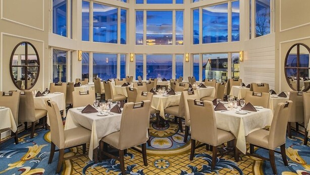 A large, well-lit dining area with multiple tables and chairs, large windows offering a view of the outside, and a decorative floor pattern.