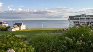 A coastal property with a view of the sea, featuring a large white house with a balcony, a smaller building, and a well-maintained lawn.