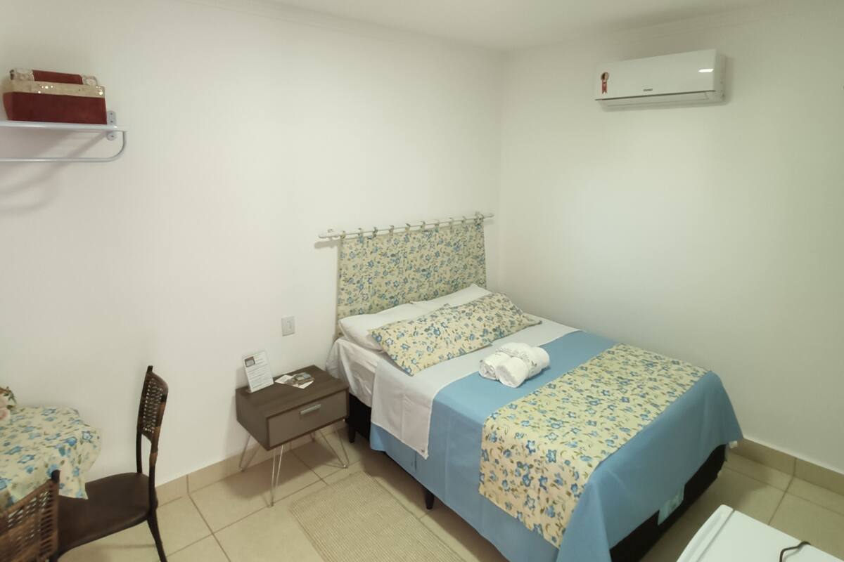 Una habitación pequeña y limpia con cama, escritorio, silla, mesita de noche, aire acondicionado de pared y una estantería con una caja.