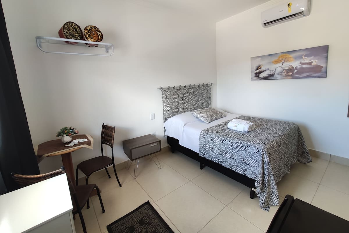 Un dormitorio ordenado con una cama, una silla, una mesita, una estantería con adornos y un cuadro en la pared.