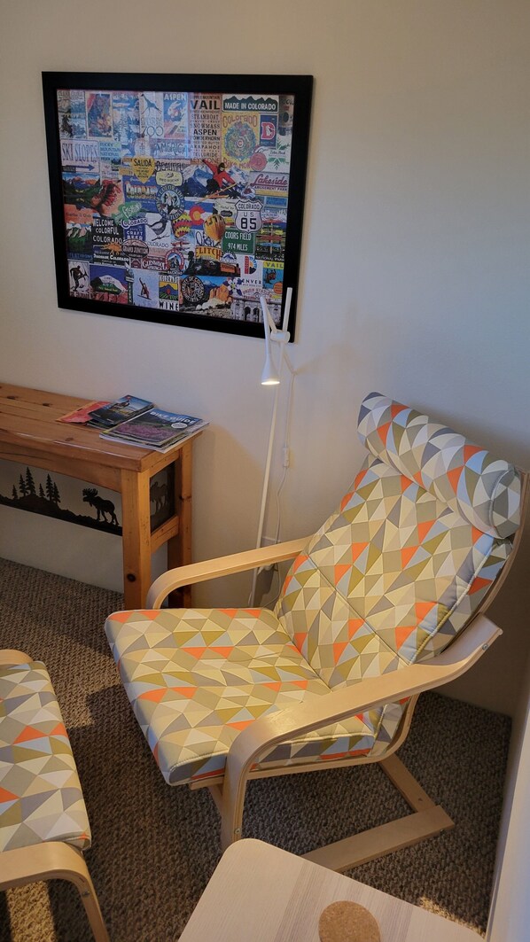 Una habitación con un televisor de pared mostrando un cartel con motivos de viaje, una mesa de centro de madera con revistas y un sillón estampado.