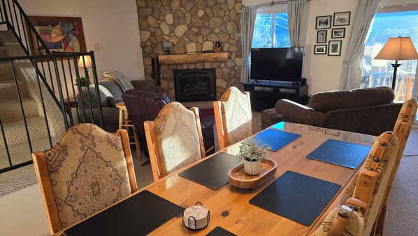 Un salon chaleureux avec une cheminée en pierre, un téléviseur et un coin repas agrémenté de chaises à motifs et d’une table en bois.