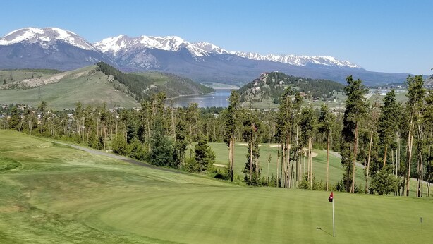 Ein Golfplatz mit Fahnenstange, umgeben von hohen Kiefern und einem Hintergrund aus schneebedeckten Bergen.