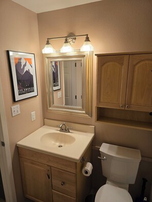 Une salle de bain avec un lavabo, un miroir et des toilettes. Un tableau encadré est accroché au mur et un luminaire se trouve au-dessus du miroir.