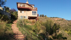 A two-story house with a balcony, a staircase leading up to it, and a hillside in the background.
