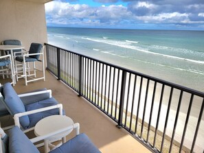 Un balcon avec des chaises blanches et une table, donnant sur une plage où l’on peut voir les vagues.