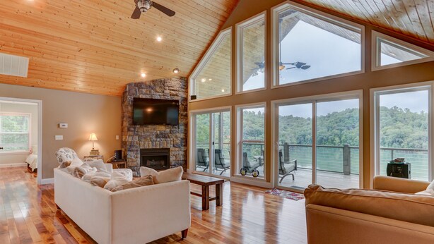 A spacious living room with a stone fireplace, large windows offering a view of the outdoors, and a ceiling fan. - Waterfront, 2025 Tram, 2023 Dock, Sleeps 14, 3BR, 3.5 Bath, Wifi @ House & Dock (Jacksboro)