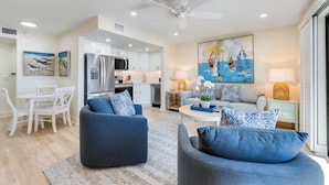 Una sala de estar moderna con un sofá y sillones, un comedor con una mesa y sillas blancas, y una cocina con gabinetes blancos y un refrigerador.