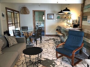 Ein Wohnzimmer mit einer Couch, einem Couchtisch, einem blauen Sessel, einem Esstisch mit Stühlen und einer Küchenzeile mit einer Bar.