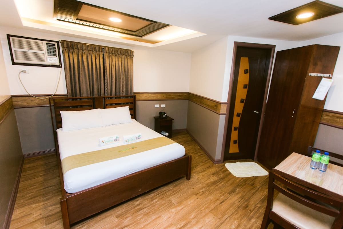 Une chambre d’hôtel avec un lit en bois, un bureau avec une chaise, une armoire, une table de chevet, un climatiseur et une fenêtre avec des rideaux.