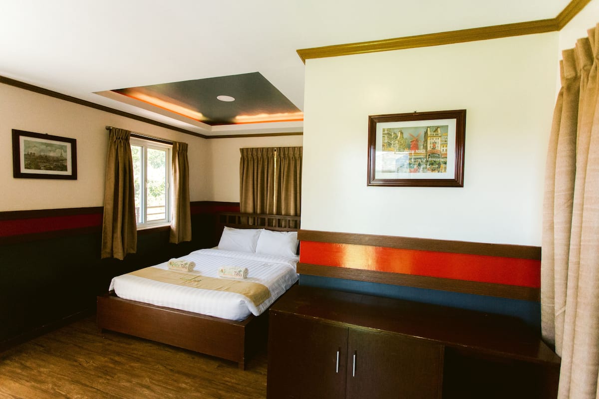 Une chambre d’hôtel avec un lit en bois, une tête de lit, une armoire, une fenêtre avec des rideaux et un tableau encadré au mur.