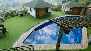 Un espace piscine bordé de pierres, entouré d’une pelouse verdoyante et de deux petits bâtiments aux toits gris.