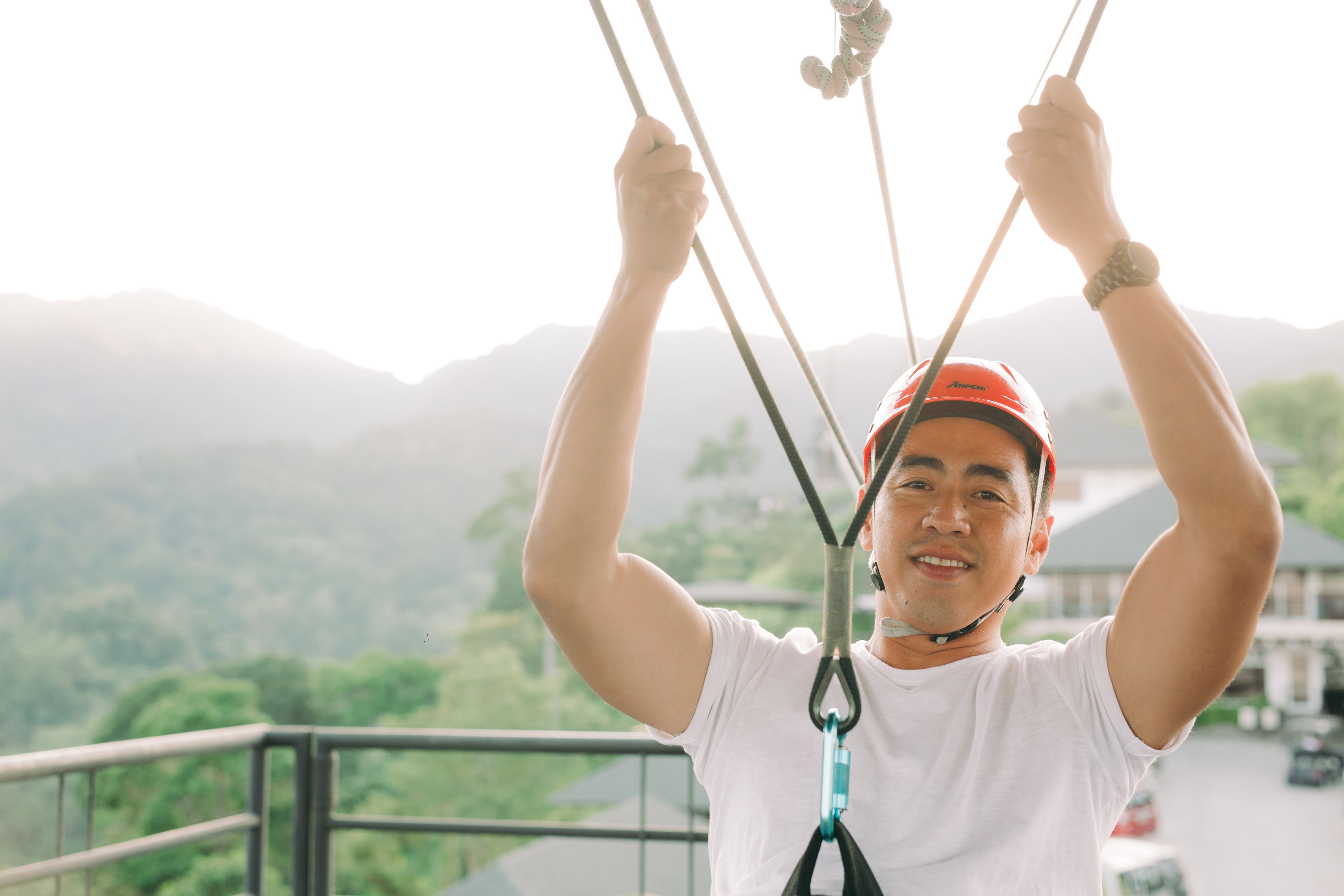 A person with a harness attached to a rope system, holding onto the ropes with both hands, with a mountainous landscape in the background.