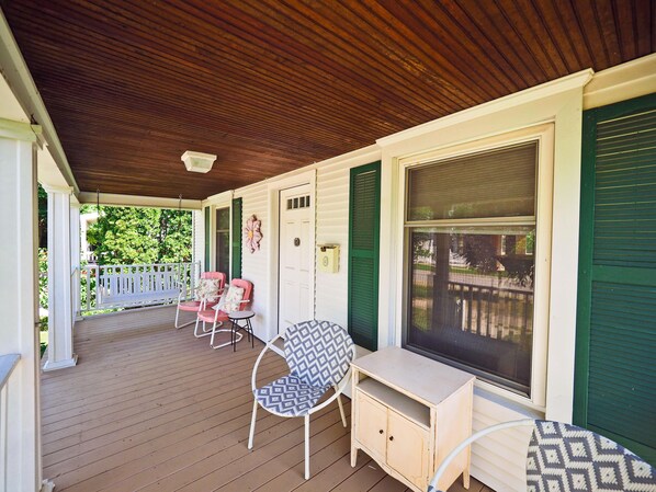 A covered porch with a wooden ceiling, white walls, green shutters, and a white door. There are two rocking chairs and a small table with a bench.