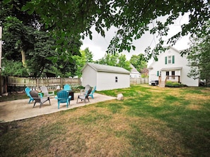 Relax in the serene backyard with Adirondack chairs and a patio table, surrounded by lush trees and a well-manicured lawn.