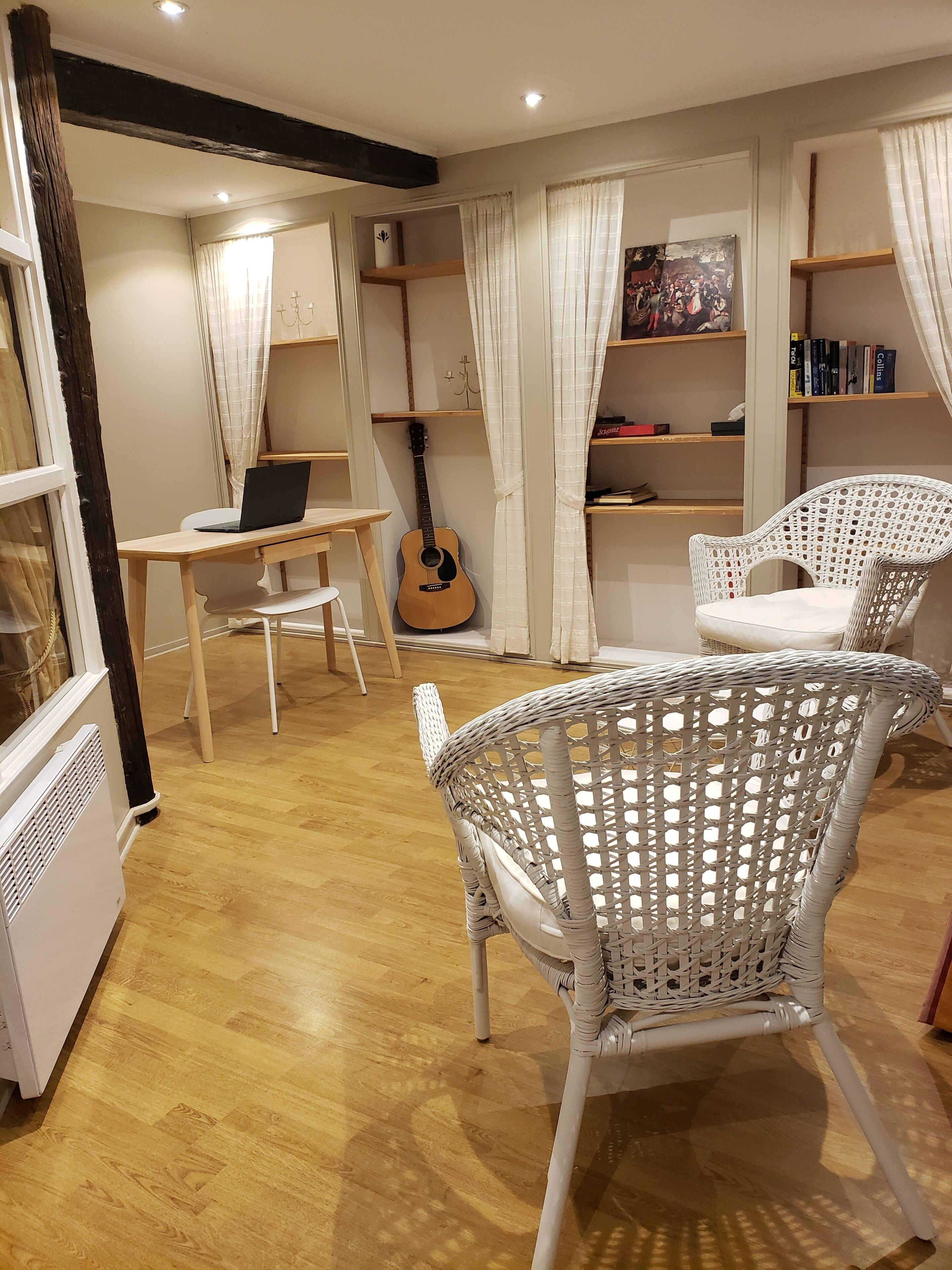 Ein gemütliches Zimmer mit Holzboden, weißen Korbstühlen, einer Gitarre, einem Laptop auf einem Tisch und Bücherregalen.