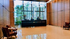 A modern reception area with a wooden reception desk, seating, and a large window with a view of bamboo plants. - Komune Living (Kuala Lumpur)