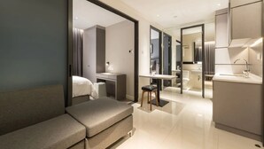 A modern hotel room with a sofa, a desk, a small stool, a vanity with a mirror, and a separate bathroom area. - Komune Living (Kuala Lumpur)