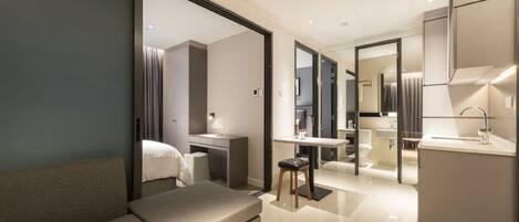 A modern hotel room with a sofa, a desk, a small stool, a vanity with a mirror, and a separate bathroom area.