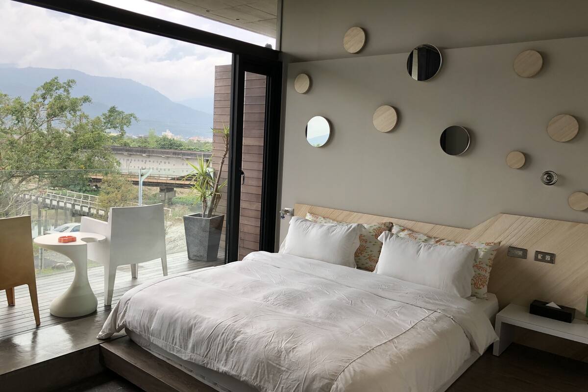 A modern bedroom with a large bed, a view of mountains, and a balcony with a table and chairs.