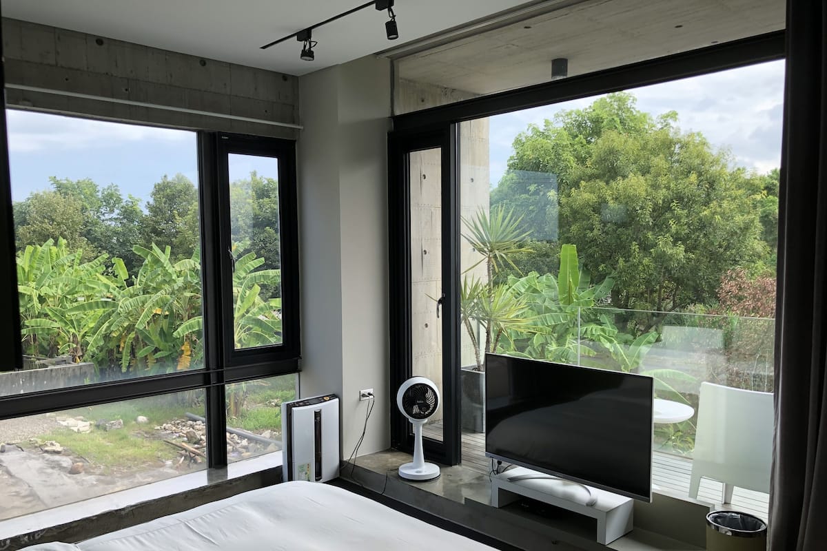 A modern bedroom with large windows, a bed, a TV, and a fan.