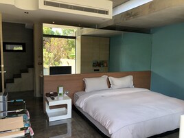 Quarto de hotel moderno com uma cama grande, uma mesa de cabeceira com controle remoto, uma escrivaninha e uma cadeira. Há uma janela com vista para árvores e uma escada que leva a outro andar.