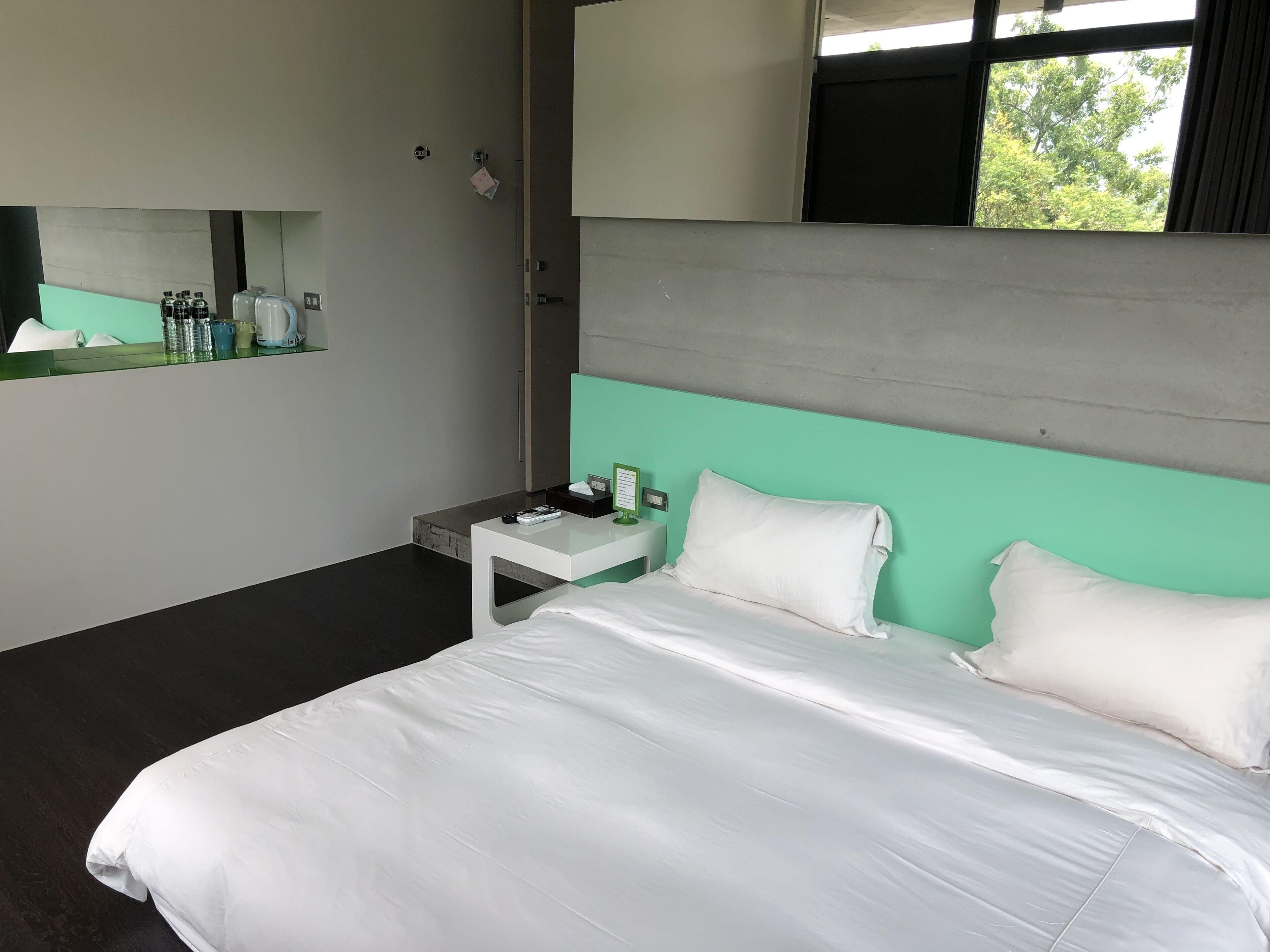 A modern hotel room with a large bed, a bedside table with a lamp, and a window with a view of trees.