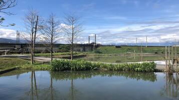 A serene outdoor area with a calm pond, trees, and a distant view of a bridge and industrial structures.