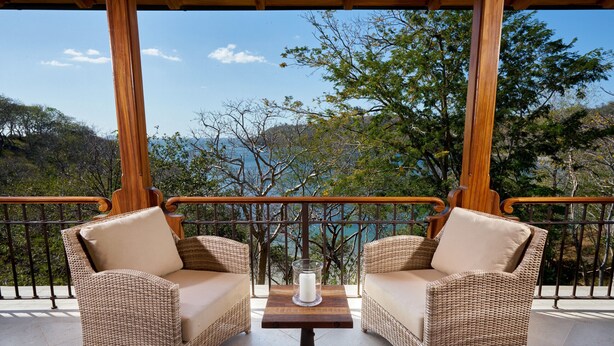 Un balcón con mobiliario de mimbre, una mesa con una vela y vistas a árboles y colinas.