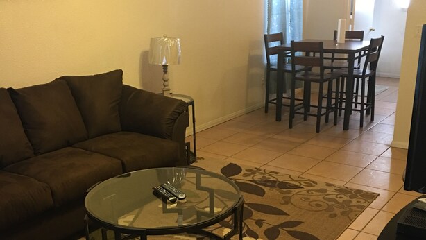 Un salon avec un canapé marron, une table basse en verre, une lampe, une table à manger avec des chaises et un tapis à motifs.