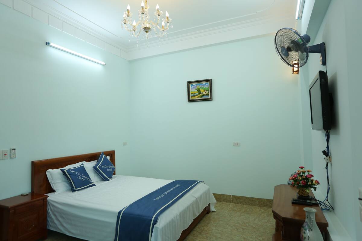 A hotel room with a bed, bedside table, nightstand, wall-mounted TV, ceiling fan, chandelier, and a painting on the wall.
