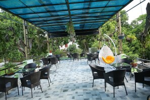 An outdoor patio with black wicker chairs and tables, covered by a blue canopy, surrounded by greenery.