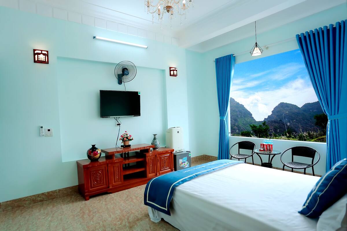 A bedroom with a bed, a TV, a fan, a small table with chairs, and a view of mountains.