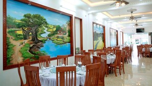 A dining area with tables set for service, large landscape paintings on the walls, and ceiling fans. - TamCoc Green field Hostel (Nam Hoa Lu)