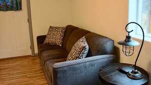 Quarto com sofá cinzento, almofadas estampadas, mesa redonda pequena com abajur e chão de madeira.