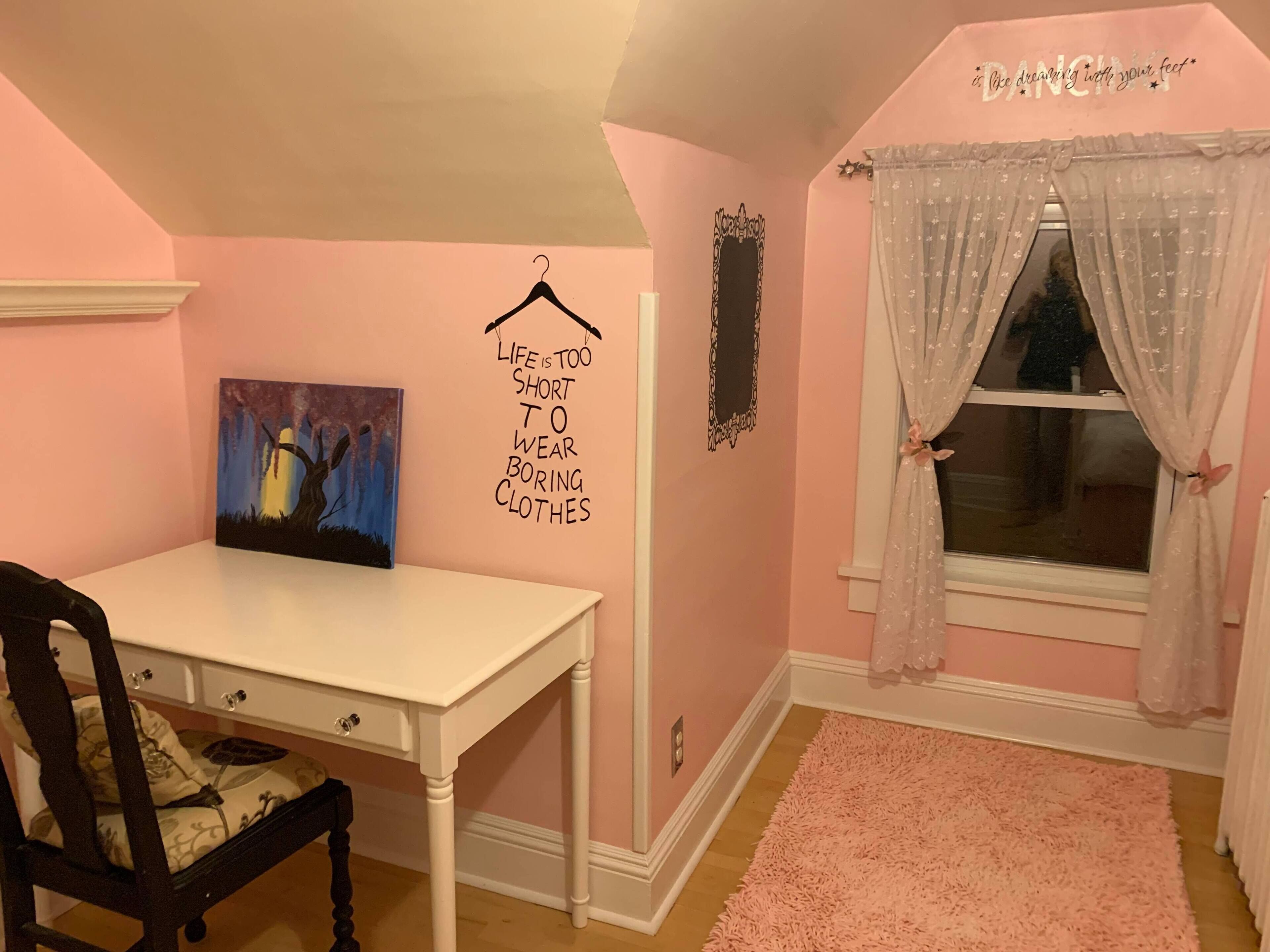 A room with a white table, a chair, a pink wall, a window with sheer curtains, and a motivational quote on the wall.