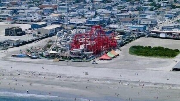 Vue aérienne d’une plage avec une grande structure rouge, qui pourrait être une jetée ou un quai, et un paysage urbain en arrière-plan.