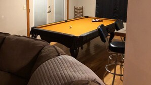 Ein Raum mit Billardtisch, einem Sessel, einem Barhocker und einer Deckenleuchte.
