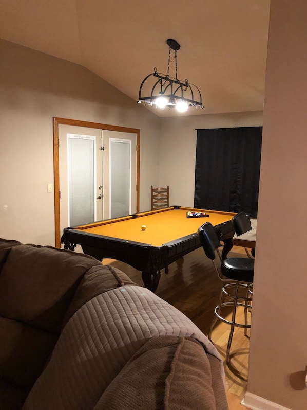 Une pièce comprenant une table de billard, une chaise, un tabouret de bar et un luminaire fixé au plafond.