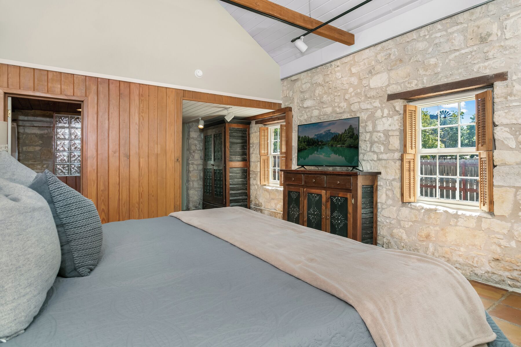 Une chambre à coucher avec un lit, des murs lambrés en bois, un mur en pierre, un téléviseur posé sur un meuble et une fenêtre avec des volets.