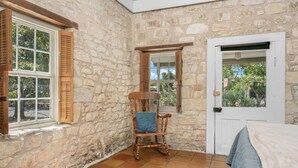 Una habitación con paredes de piedra, una silla de madera con cojín, una cama con manta azul y una puerta con ventana.