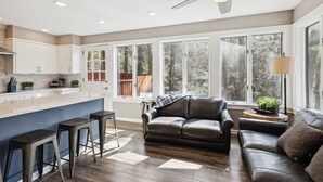 Una cocina moderna con una isla central, taburetes de bar, un sofá de cuero y ventanas amplias con vistas al exterior.