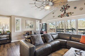 A spacious living room with a leather sectional sofa, a ceiling fan, and large windows offering a view of the outdoors. - Downtown Cabin w/Ball Court, Hot Tub, & Firepit (GATLINBURG)