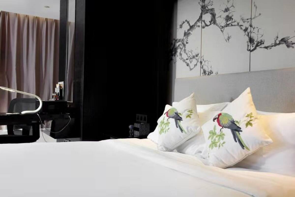 Ein Hotelzimmer mit Bett, Schreibtisch und Stuhl. Es gibt ein Fenster mit Vorhängen, eine Wand mit einem Gemälde von einem Ast und ein Kissen mit Vogelmotiv.