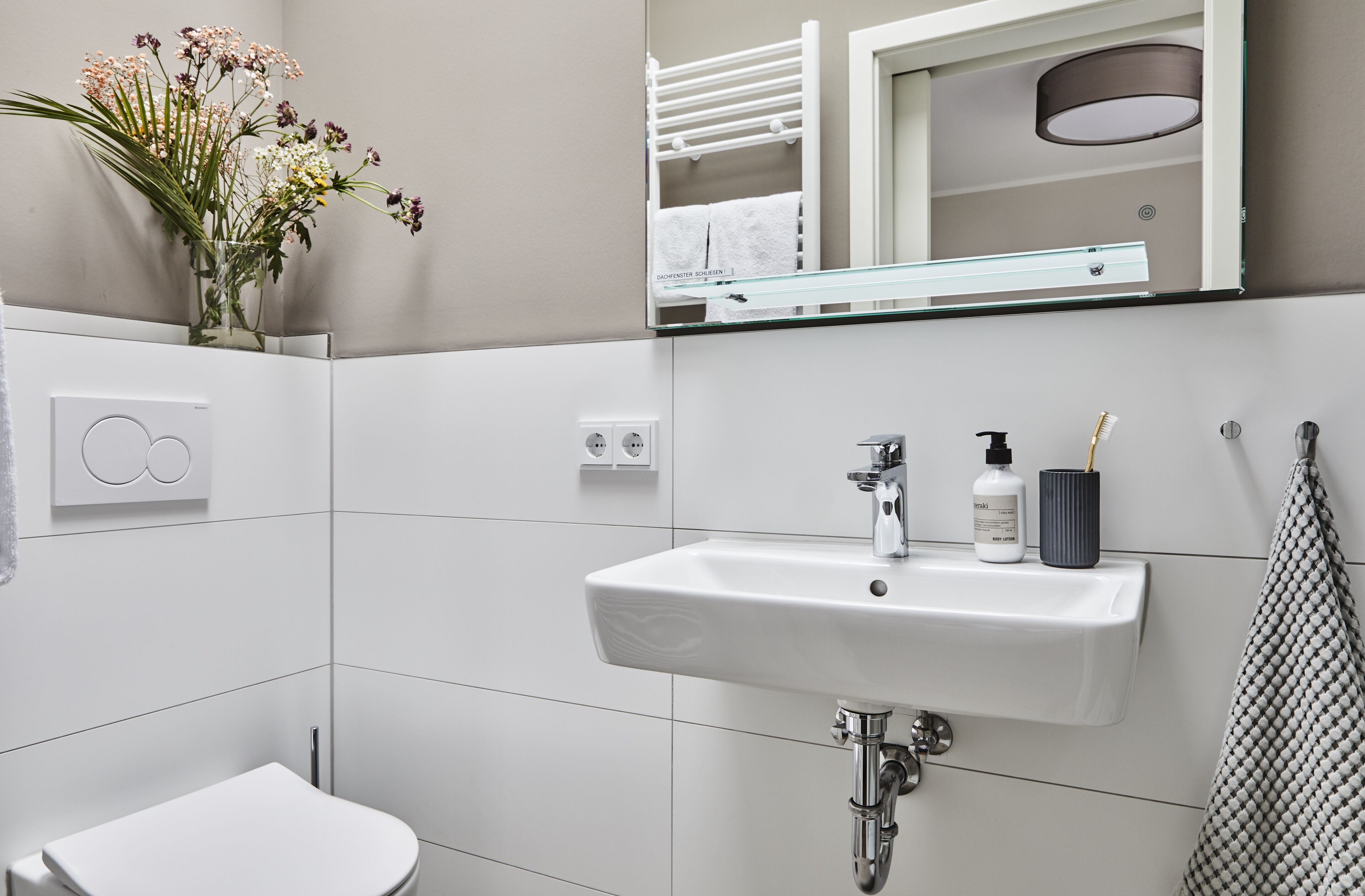 Une salle de bain avec un lavabo blanc, un miroir et des toilettes. Un vase rempli de fleurs est posé sur le mur, et une serviette est accrochée sur le côté.