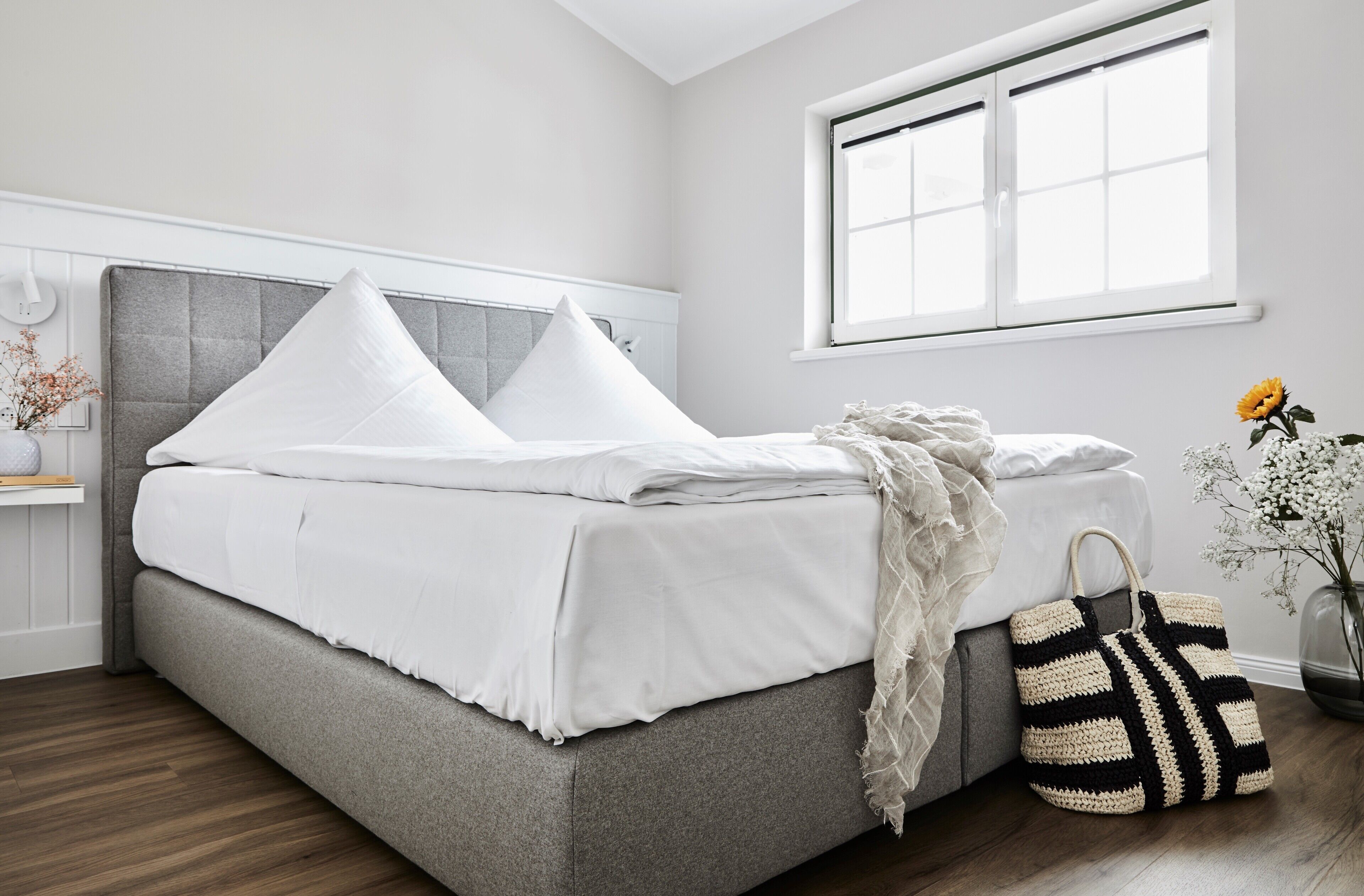 A neatly made bed with white bedding and pillows, a grey upholstered headboard, and a window with white frames.