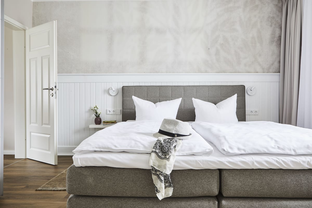 A neatly made bed with white linens and a grey headboard, a white door, and a small bedside table with a vase.